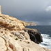 An der Mittelmeerküste östlich von Wied iż-Żurrieq - Ausblick entlang der Klippen. Links ist der Sciuta Tower (aka Ta' Xuta tower) zu sehen, im Hintergrund gehen Regenschauer nieder.