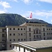 Kaserne Andermatt mit Schweizerfahne vor Bergpanorama.