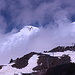 Zwischen Mir und Garabashi - Ausblick während der Fahrt mit dem Sessellift. Hinter den Wolkenfetzen zeigen sich beide Elbrus-Gipfel. In der Vulkanlandschaft im Vordergrund sind nebenbei neue Stützen zu sehen, offenbar für eine weitere Verlängerung der Seilbahn. Foto vom 17.07.2015.