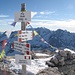Der Wegweiser auf dem Gipfel ist mit tibetischen Fähnchen behängt .... im Hintergrund der höchste Dolomitenberg, die Marmolata (Punta Penia, 3343m) und rechts daneben der für Durchschnittsbergsteiger unzugängliche Gran Vernel (3210m)