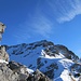 Das Gipfelkreuz taucht auf. Ein Stück Drahtseil, das zu sehen ist, gibt Aufschluss darüber, dass es rechts am Felskopf vorbei über das Schneeband gehen wird
