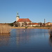 Most - Blick über den Teich zur Kirche Mariä Himmelfahrt. Diese wurde 1975 von ihrem ehemaligen Standort um 841 m verschoben und befindet sich nun an der letzten erhaltenen Straße des "alten" Most. Auf dem Wasser tummeln sich derweil einige Schwäne und Blässrallen.