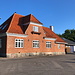 Rø - Straßenseitiger Blick auf das ehemalige Bahnhofsgebäude. Hinten ist auch der frühere Güterschuppen zu sehen. Auf Bahnhofsplänen der Bornholmer Eisenbahnen sind i. d. R. die Bezeichnungen "Hovedbygning" (Hauptgebäude) bzw. "Varehus" (Warenhaus) eingetragen.