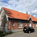 Aakirkeby - Straßenseitiger Blick auf das ehemalige Bahnhofsgebäude, das sich bei km 17,4 und damit etwa in der Mitte der ehemaligen "[https://da.wikipedia.org/wiki/Nex%C3%B8banen Nexøbanen]" (Rønne - Nexø) befindet.